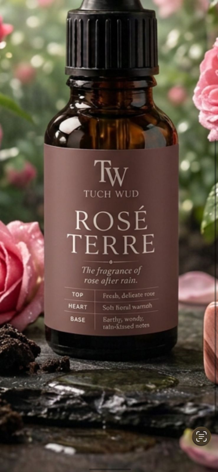 A bottle of TW Tuch Wld Rosé Terre fragrance oil, an amber glass dropper bottle with a dark brown label, set against a blurred background of pink roses and green foliage.